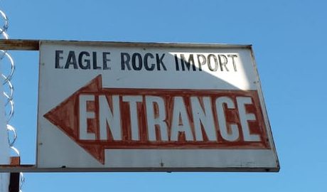 A weathered white sign on a bracket reading Eagle Rock Import with a red arrow pointing to the entrance