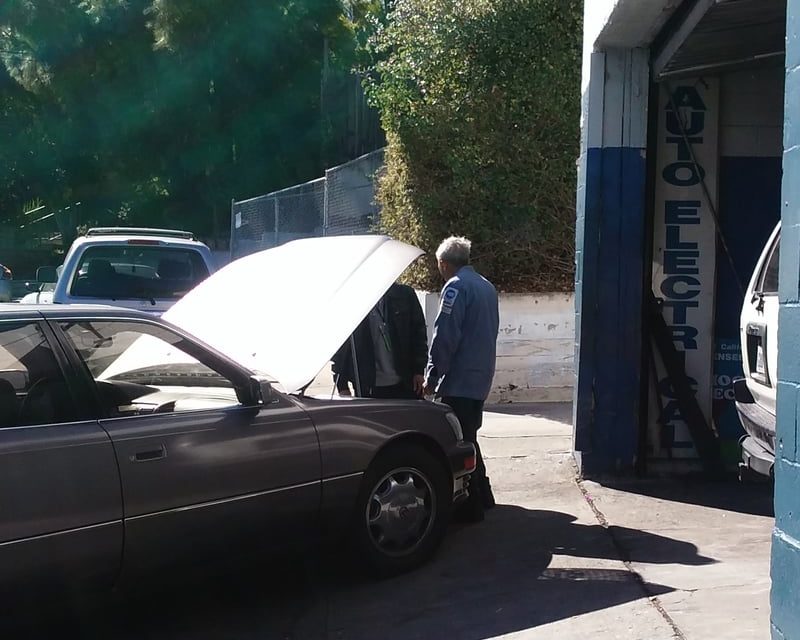 A mechanic in a blue work shirt looking under the raised hood of a car with its owner, beside the open bay marked Auto Electrical